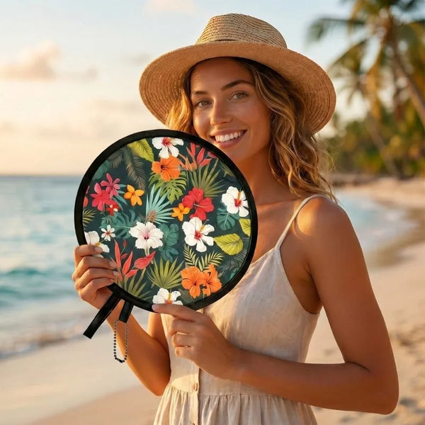 A smiling woman wearing a straw hat holds an open floral foldable pocket fan outdoors near the sea.