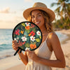 A smiling woman wearing a straw hat holds an open floral foldable pocket fan outdoors near the sea.