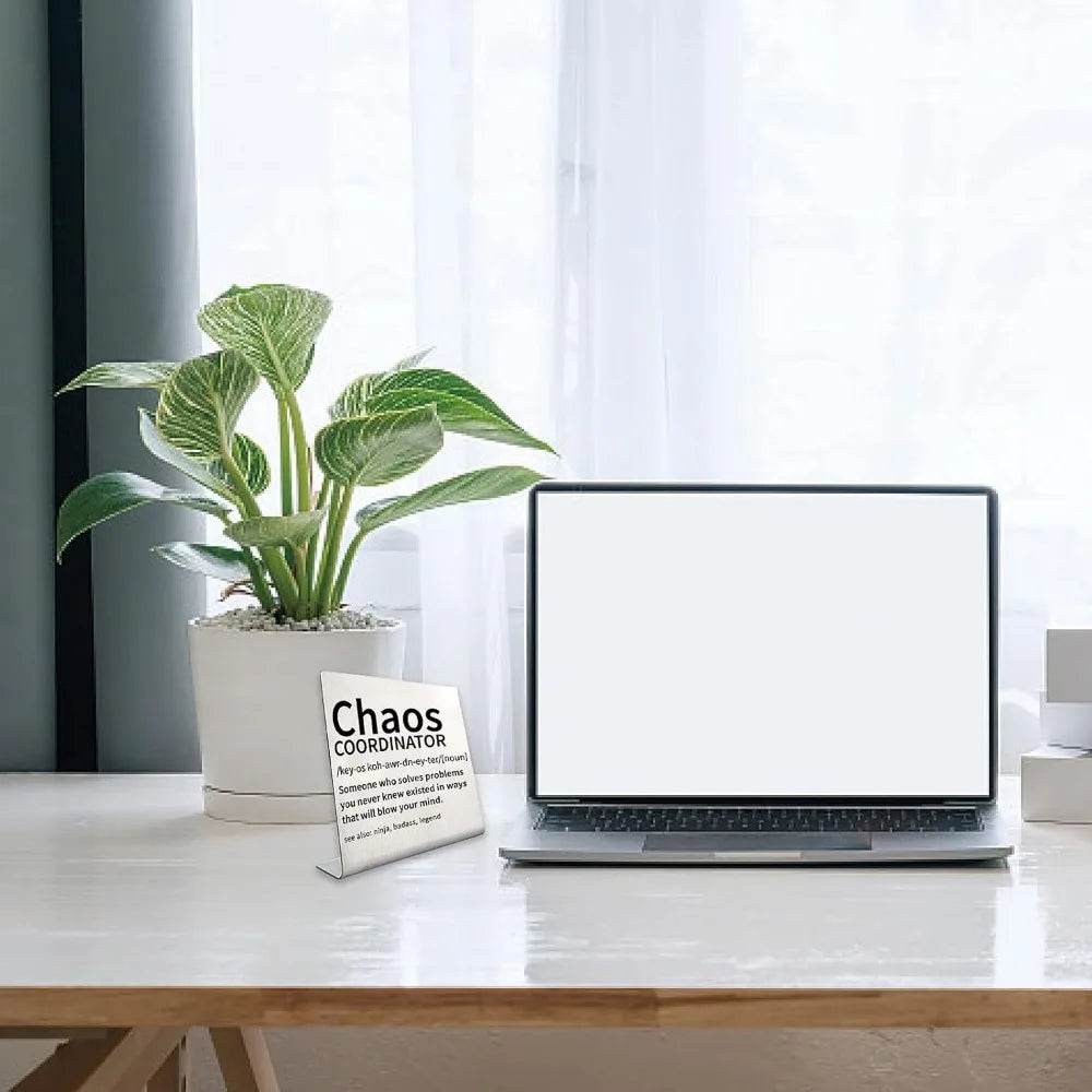 Una laptop colocada en un escritorio blanco junto a una planta en maceta y un letrero etiquetado como Coordinador del Caos.