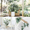 A bundle of eucalyptus leaves in a woven basket set on a wooden table with greenery and white flowers.