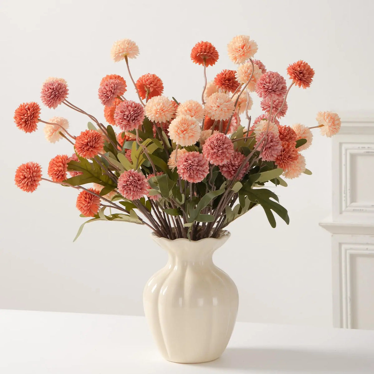 A bouquet of artificial chrysanthemum pompon flowers in shades of pink and orange arranged in a white vase.