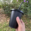 A hand holds a black plastic cold cup with a lid and straw outdoors amidst green foliage.
