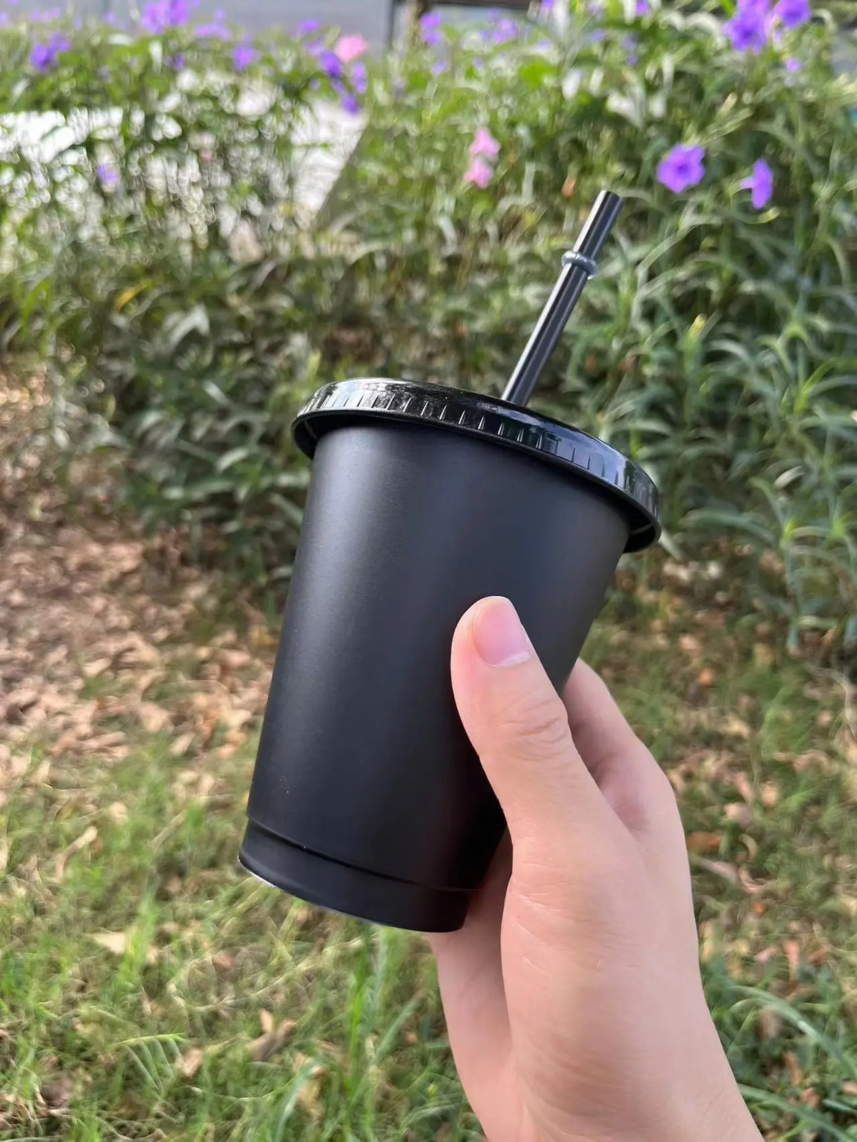 A hand holds a black plastic cold cup with a lid and straw outdoors amidst green foliage.