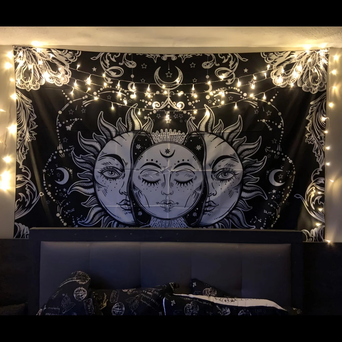 A dark tapestry featuring celestial sun and moon faces with zodiac art illuminated by string lights.