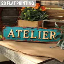 A vintage industrial metal sign with the word ATELIER in golden lettering on a turquoise distressed background.