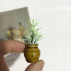 A hand holds a small, olive-green ceramic vase containing artificial white and green sprigs.
