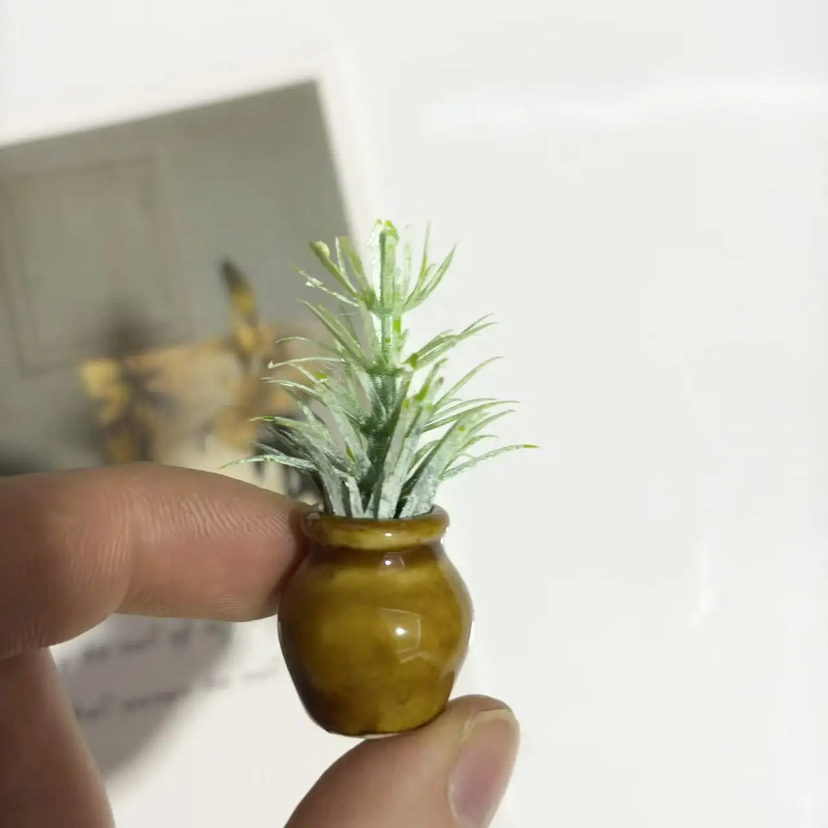 A hand holds a small, olive-green ceramic vase containing artificial white and green sprigs.