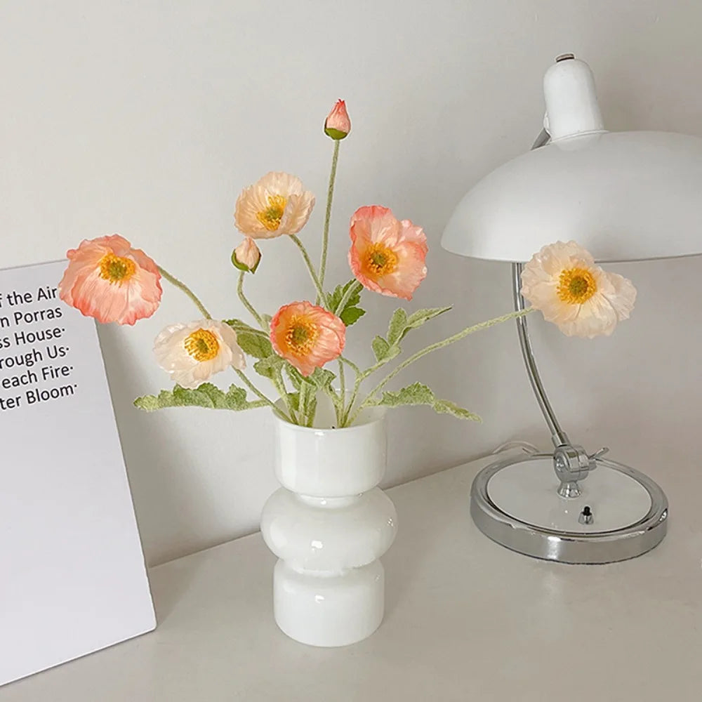 A white ceramic flower vase holds several delicate glass flowers on a light gray table next to a lamp.