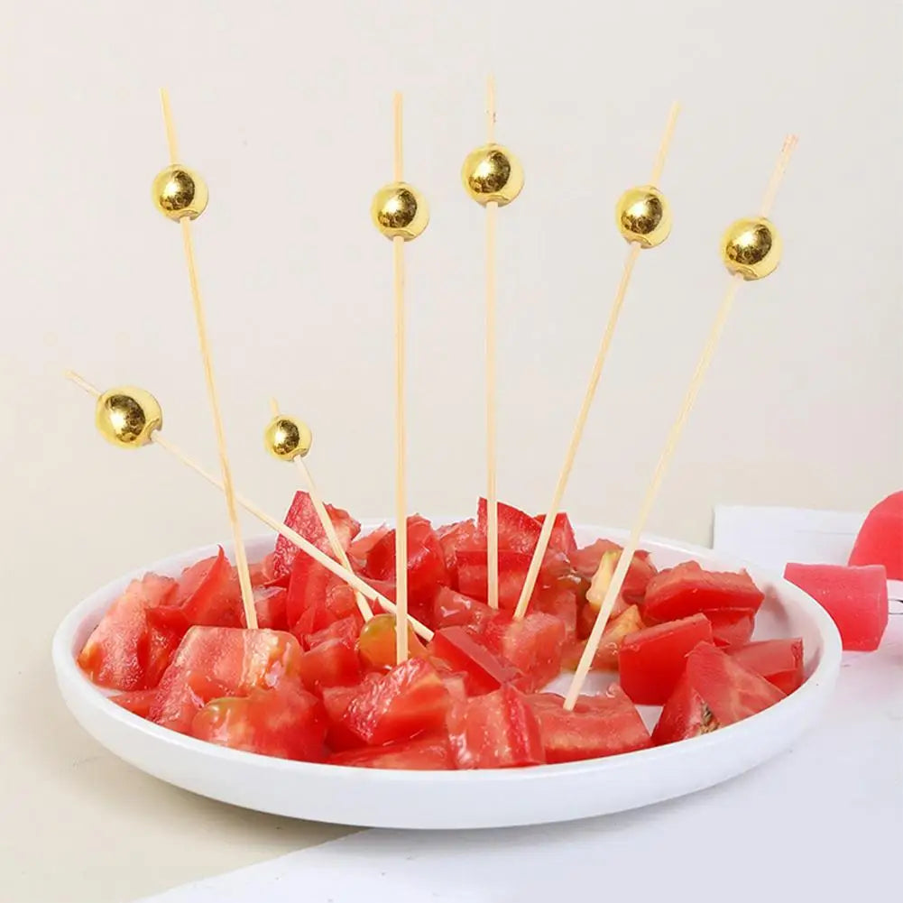 A white plate holds sliced tomatoes skewered with gold accent bamboo sticks for appetizers.