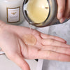 A hand pours a clear wax from a glass jar into the palm of another hand for a hand-poured natural soy candle.