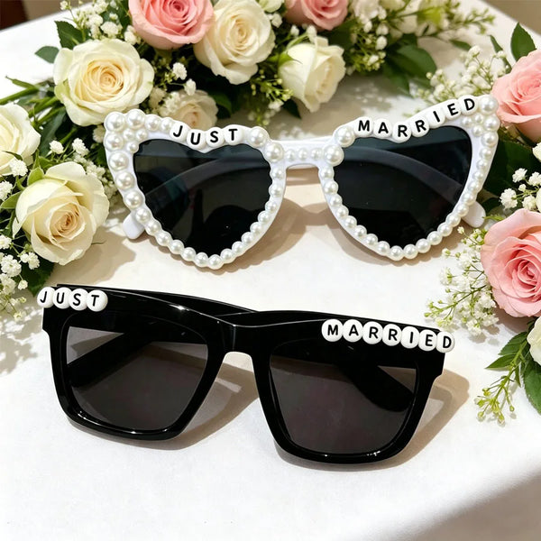 A set of black sunglasses decorated with pearl borders and 'just married' text, displayed on a white table surrounded by pink