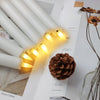 A set of six flameless LED taper candles glowing yellow on a white surface next to a pine cone.