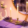 A tall white candle in a brass candlestick holder sits on a purple cloth next to lavender flowers.
