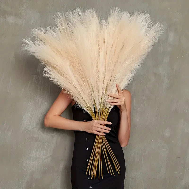 A woman holds a large, fluffy beige pampas grass stem against a gray background.