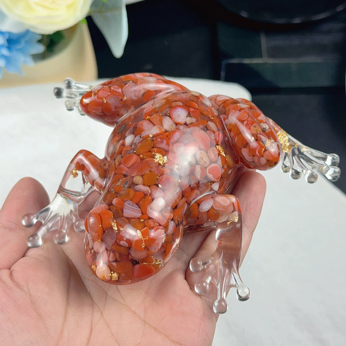 A hand holds a clear resin craft decoration shaped like an orange and pink frog with gold accents.
