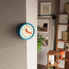 A modern resin wall clock with a blue face and orange hands is mounted on a light gray wall.