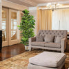 A large artificial fiddle leaf fig tree in a white pot stands next to a beige upholstered sofa in a well-lit living room.