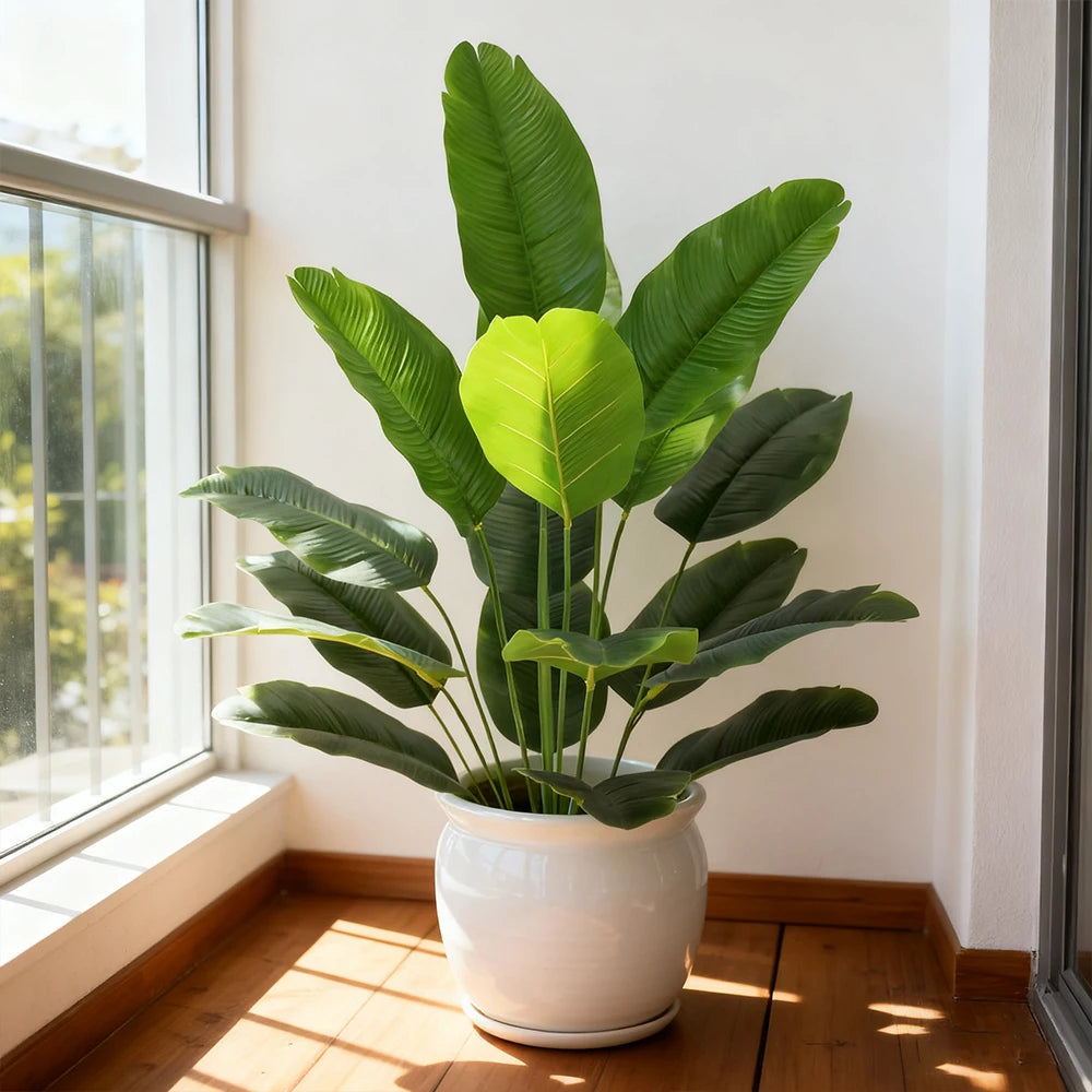 A vibrant green artificial Bird of Paradise plant with large leaves in a white pot sits on a wooden floor next to a window.