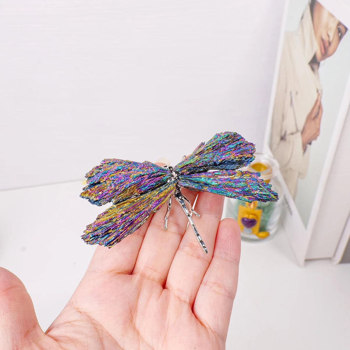 A hand holds a sparkling natural crystal tourmaline dragonfly figurine with iridescent blue and purple colors.