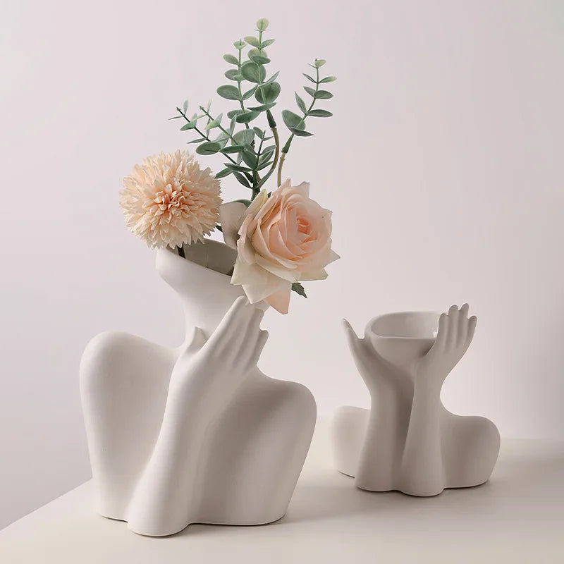 A white ceramic figural vase holding dried greenery and a pale pink rose.