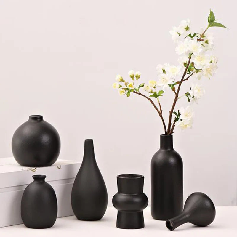 A collection of matte black ceramic vases and a branch with white blossoms on a white surface.