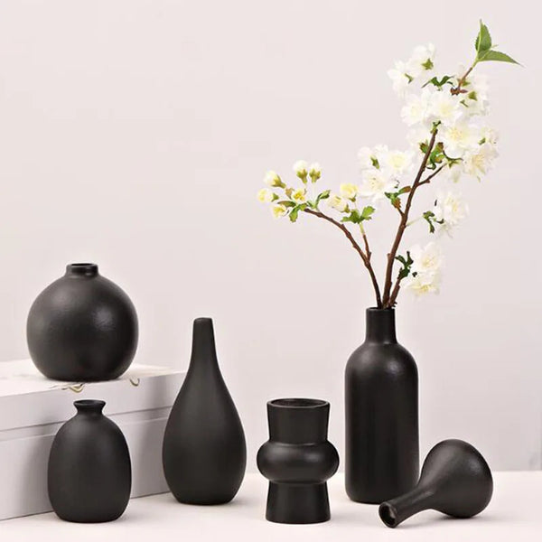 A collection of matte black ceramic vases and a branch with white blossoms on a white surface.