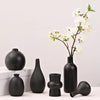 A collection of matte black ceramic vases and a branch with white blossoms on a white surface.