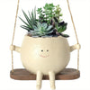 A resin succulent planter with a smiling face and a green plant arrangement hangs from a rope.