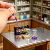 A hand uses a pipette to transfer purple liquid into small glass vials on a metal table in a laboratory setting.
