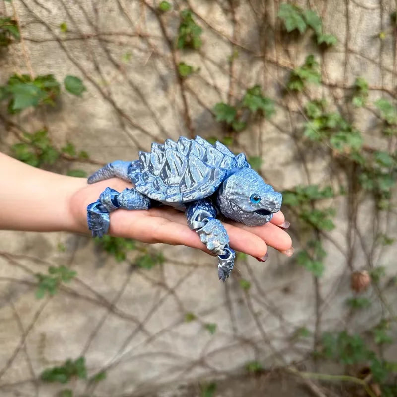 A hand is holding a blue and gray 3D printed alligator snapping turtle figurine with articulated joints against a blurred gre