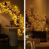 A close-up of a decorative tree branch illuminated by warm LED vine string lights against a neutral wall.