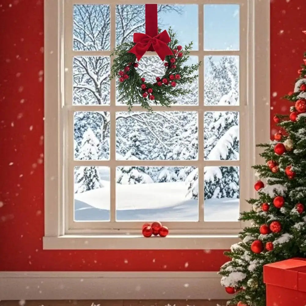 An artificial Christmas wreath with red berries and a velvet bow hangs in a window frame against a snowy winter background.
