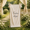 A cream linen banner with custom design text displayed outdoors on a wooden stand surrounded by greenery and white flowers.