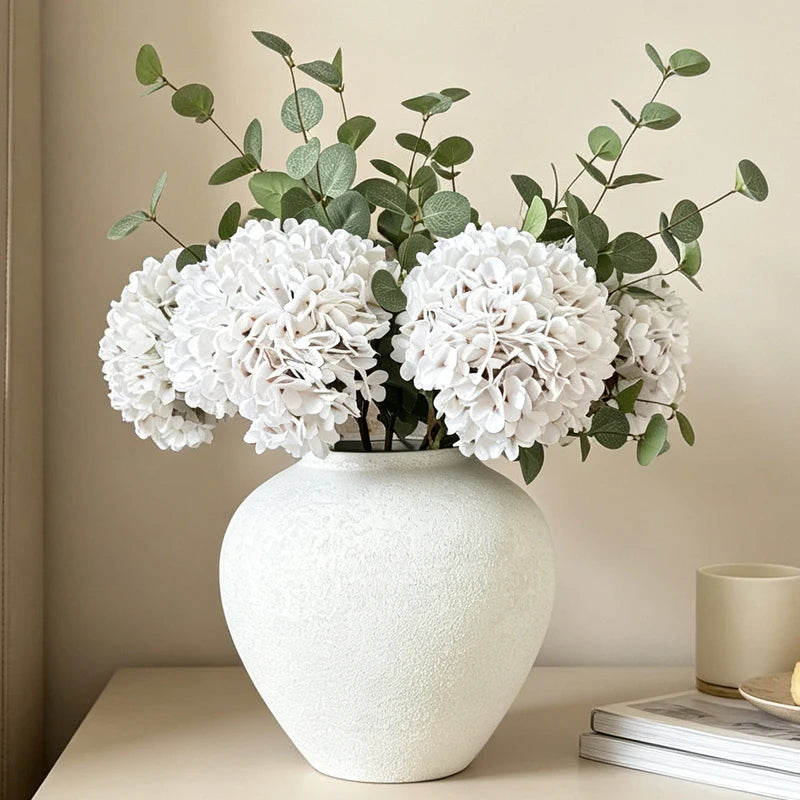 A rustic textured round white vase filled with a bouquet of white hydrangeas and green foliage.
