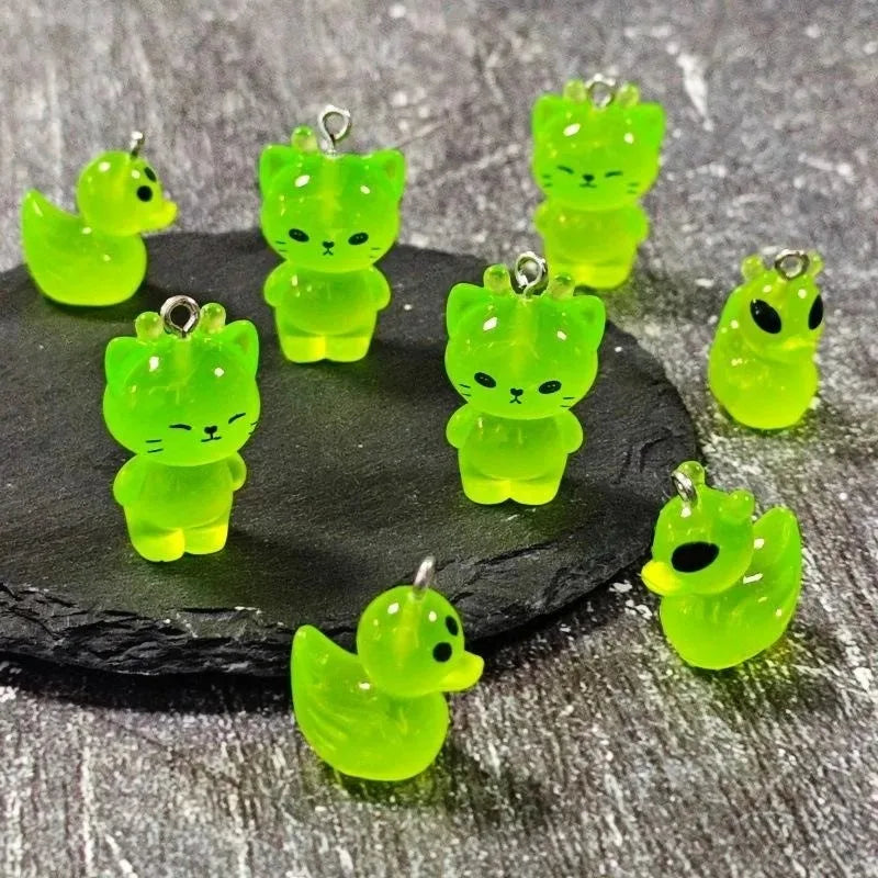A collection of bright green glow-in-the-dark cartoon alien kitty duck resin charms displayed on a dark surface.