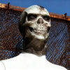 A realistic 3D skull balaclava mask covering the face, worn against a rusty wire fence under a blue sky.