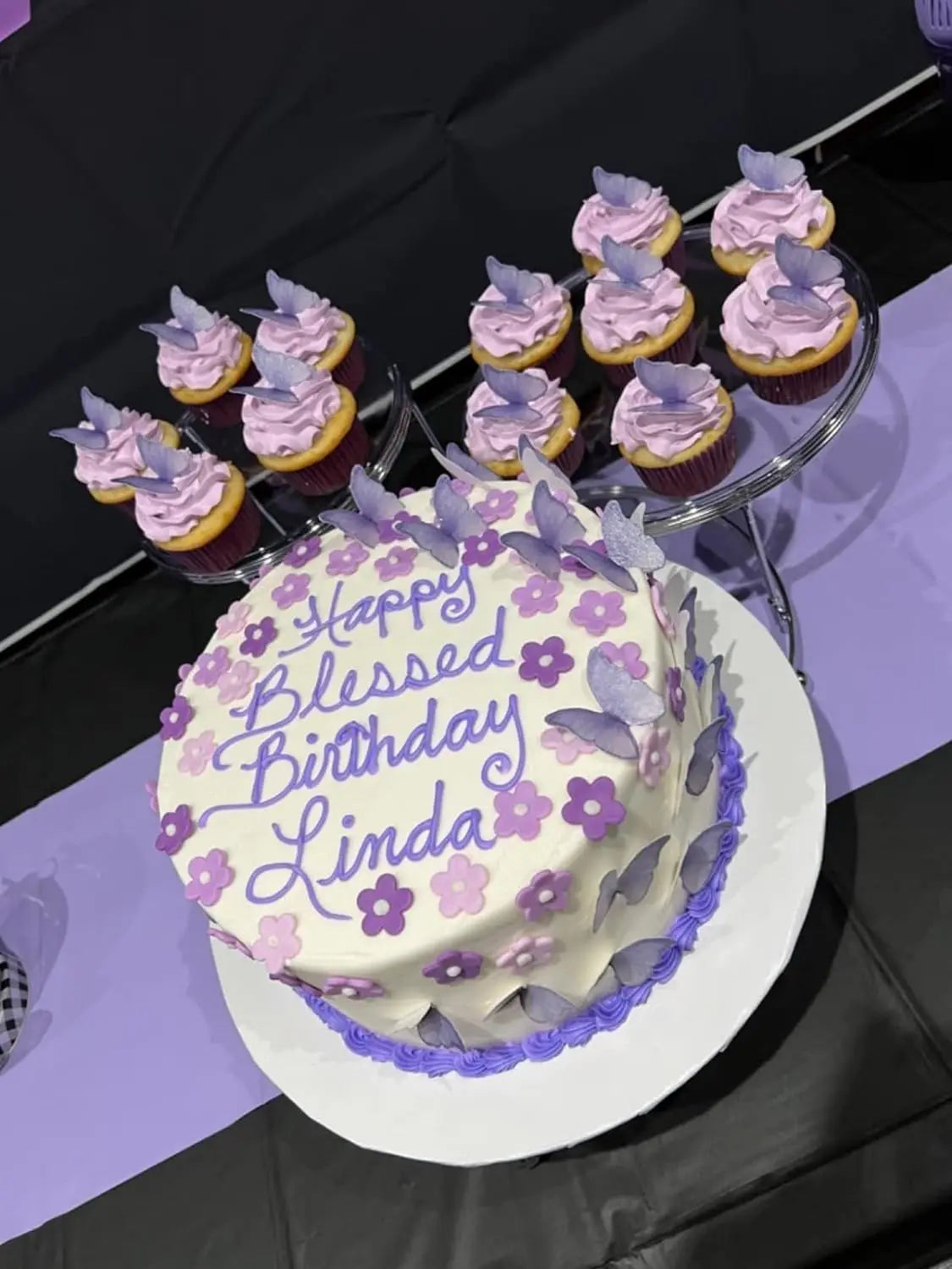 A birthday cake decorated with purple and pink flowers, surrounded by small cupcakes topped with matching butterfly decoratio