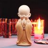 A small, light brown resin figurine of a meditating monk with hands clasped in prayer, set against a dark background with a l