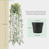 Une longue plante artificielle d'eucalyptus verte et pendante dans un pot noir sur un fond clair.
