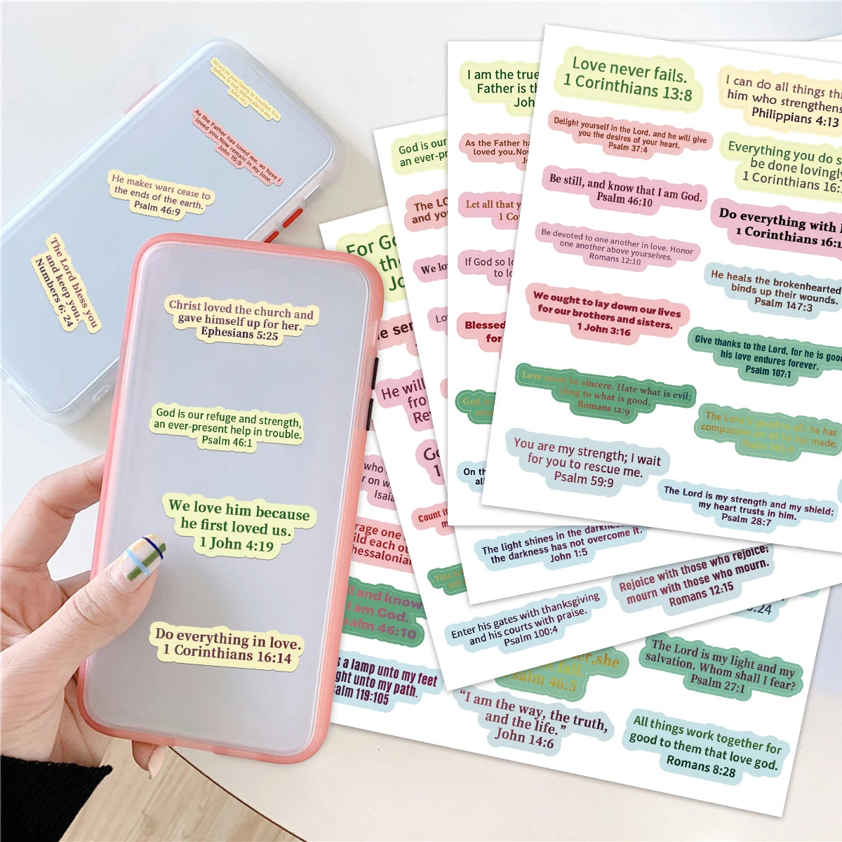 A hand holds a pink smartphone next to a stack of colorful Bible verse stickers for journals and planners.