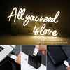 A neon sign spelling out All you need is love with warm LED lights shown alongside various charging cables and power banks.