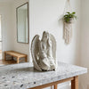 Estatua de ángel de mármol blanco con alas, exhibida en una mesa de madera contra una pared clara.