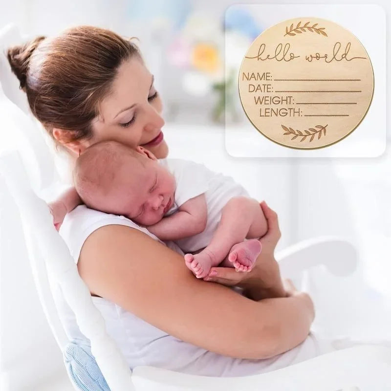 A woman holds a newborn baby while looking at a personalized wooden name announcement sign with blank lines for details.