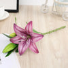 A realistic silk lily flower stem with three vibrant purple blooms and green leaves on a light wooden surface.