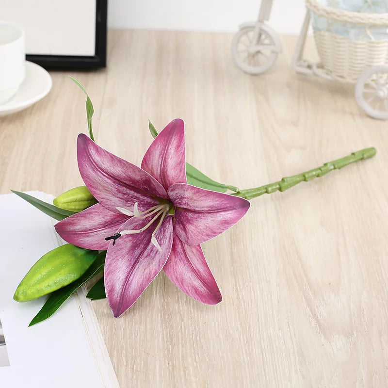 A realistic silk lily flower stem with three vibrant purple blooms and green leaves on a light wooden surface.