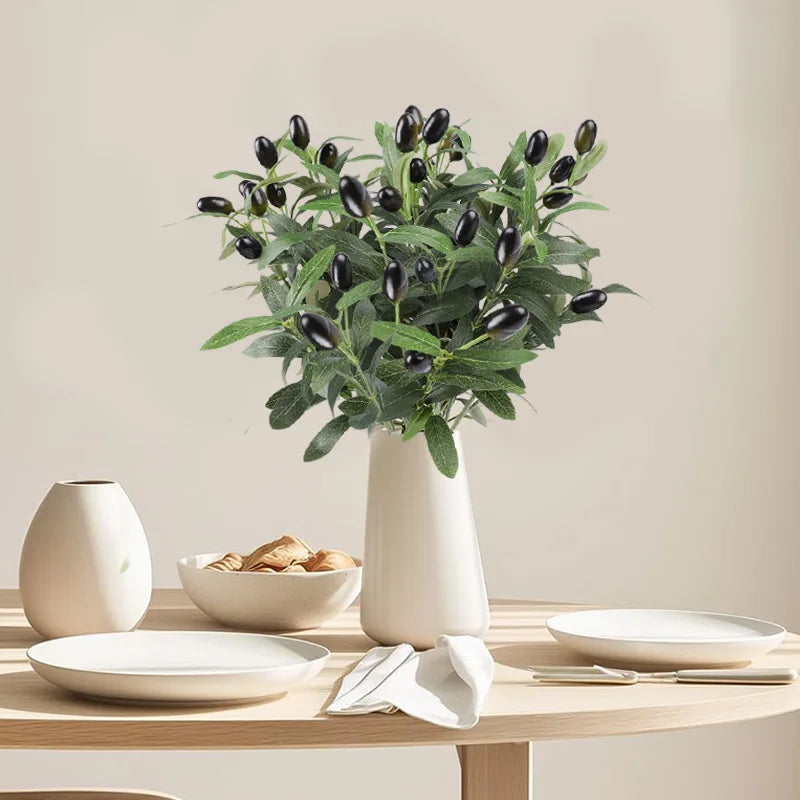 Artificial olive leaf branch in a white vase on a wooden table with plates and silverware