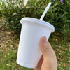 A hand holds a white plastic cold cup with a lid and straw outdoors.