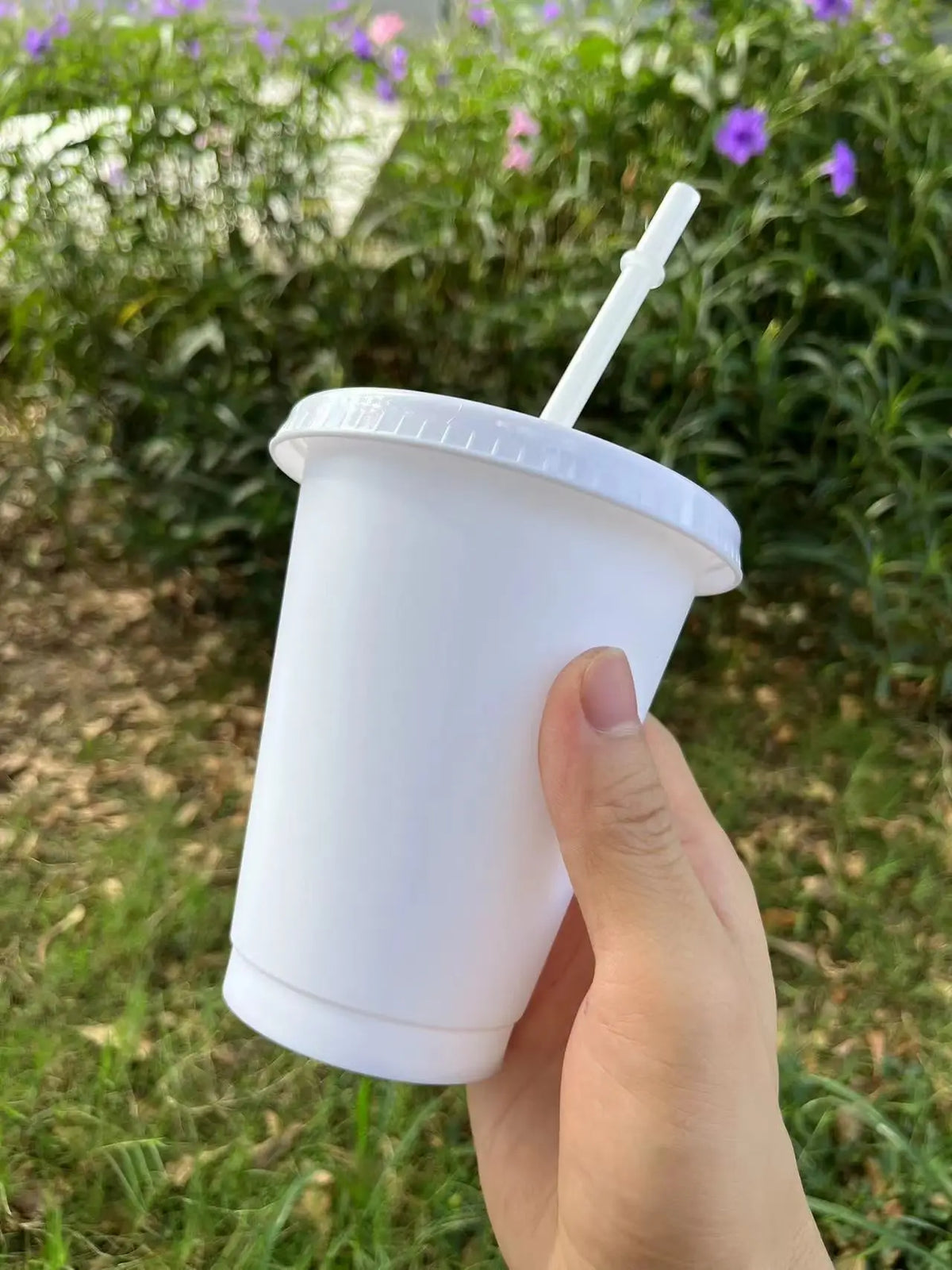 A hand holds a white plastic cold cup with a lid and straw outdoors.