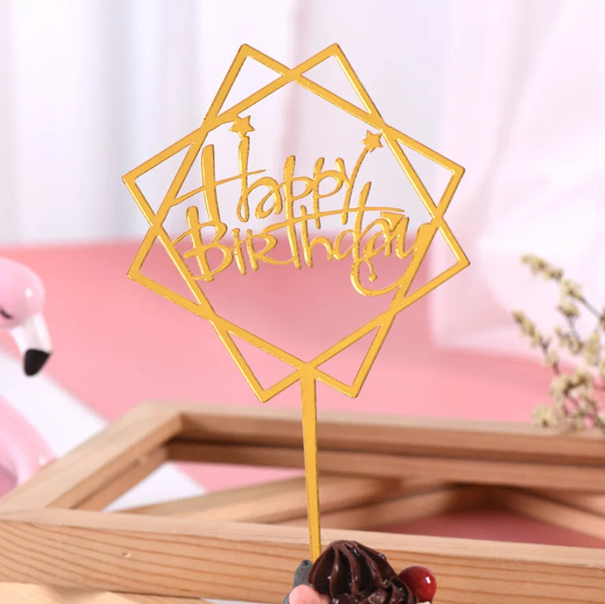 A gold acrylic happy birthday topper with a geometric shape and text on a wooden background.