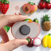 A hand holds a pink and green fruit fridge magnet with a round silver center, surrounded by various fresh fruits.